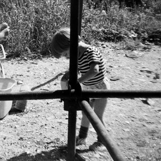 Child playing near a tub outdoors with relaxed adult in background.