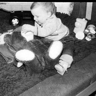 Toddler with a large teddy bear and toy tricycle on a shag rug