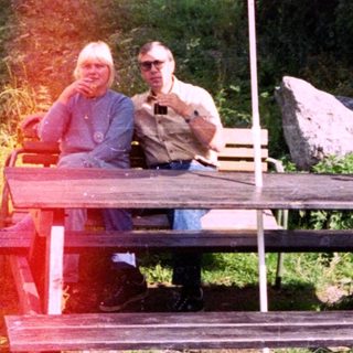 Two people relaxing under umbrella at outdoor picnic table.