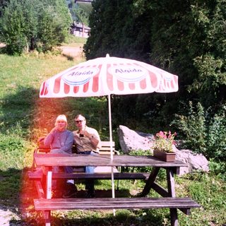Two people relaxing under umbrella at outdoor picnic table.