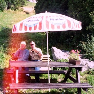 Two people relaxing under umbrella at outdoor picnic table.