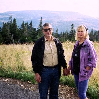 Two people standing outdoors in a scenic, forested hilly area.