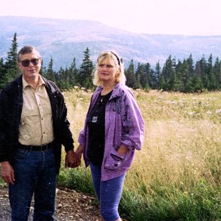 Two people standing outdoors in a scenic, forested hilly area.