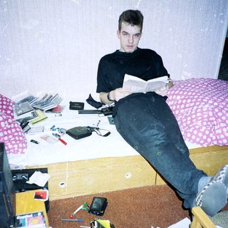 Young man reading in cluttered, disorganized bedroom.