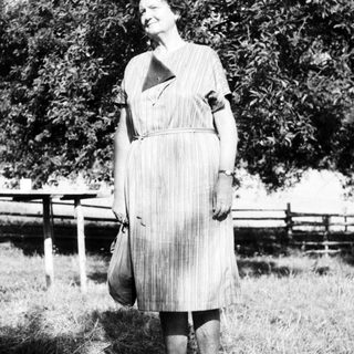 Woman in vintage dress standing outdoors, holding bag.