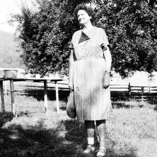 Woman in vintage dress standing outdoors, holding bag.