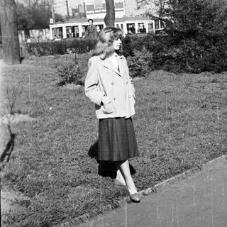 Woman in 1950s-style coat and skirt walking outdoors.