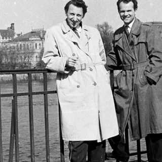 Two men posing by a bridge by water.