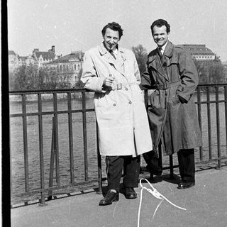 Two men posing by a bridge by water.