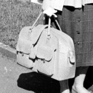 Young woman in 1960s-style coat and skirt carrying suitcase.