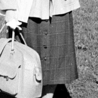 Young woman in 1960s-style coat and skirt carrying suitcase.