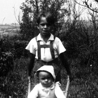 Two children in vintage clothing pushing a baby buggy outdoors.