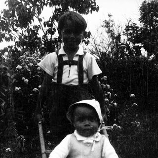 Two children posing outdoors, one standing, one seated.