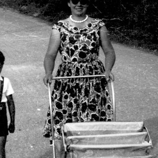 A woman pushes a stroller with a child walking beside her on a rural road.