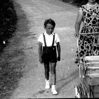 A woman pushes a stroller with a child walking beside her on a rural road.