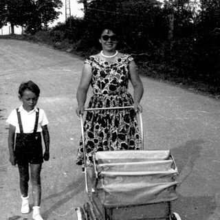 A woman pushes a stroller with a child walking beside her on a rural road.