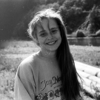 Smiling girl outdoors near water, wearing "Jay No..." shirt.