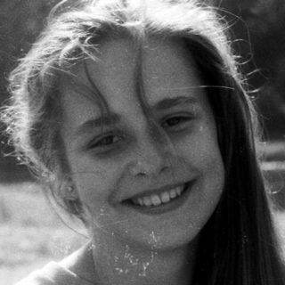 Smiling girl outdoors near water, wearing "Jay No..." shirt.
