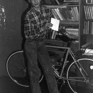 Young person holding envelope near bicycle in cluttered room.