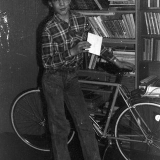 Young person holding envelope near bicycle in cluttered room.