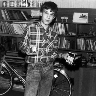Young boy holding calculator beside bicycle in book-filled room.