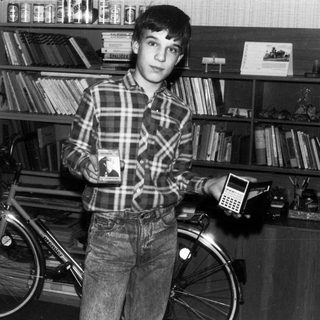Young boy holding calculator beside bicycle in book-filled room.