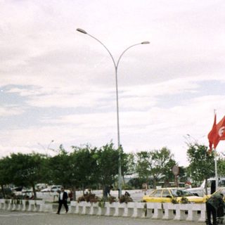 Urban street with taxis, flags, and pedestrians near a roadside.