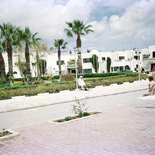 Mediterranean-style plaza with palm trees, white buildings, and people relaxing.