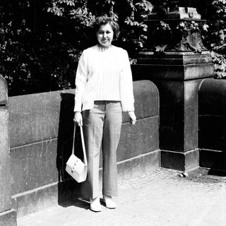 Woman standing outdoors near stone wall holding bag.