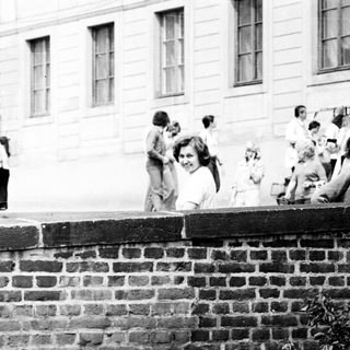 People gathered outdoors near brick wall in 1970s-style setting.