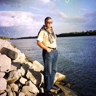 Man standing by rocky riverbank under cloudy sky.