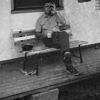Man sitting on bench outside, holding cup, wearing cap.