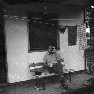Man sitting on bench outside, holding cup, wearing cap.