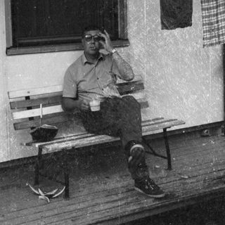 Man sitting on bench outside, holding cup, wearing cap.