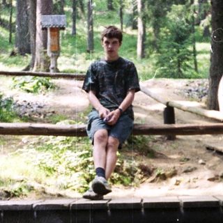 People relaxing by a wooden bridge in forested area near stream.