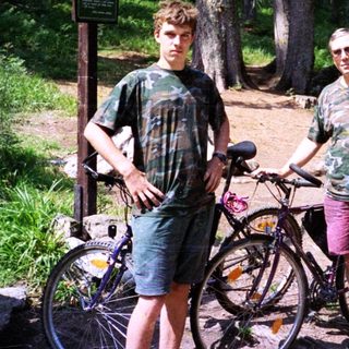 Two men posing with bicycles in a forested area.