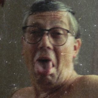 Man sitting shirtless indoors, mouth open, near a lamp.