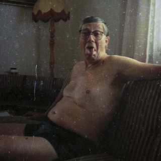 Man sitting shirtless indoors, mouth open, near a lamp.