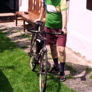 Person in cycling gear stands with bike near house.