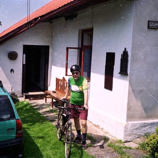 Person in cycling gear stands with bike near house.