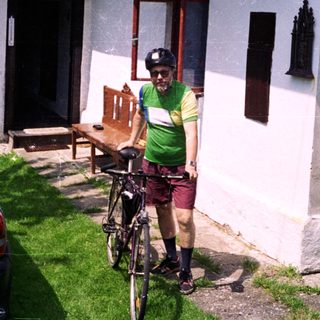 Person in cycling gear stands with bike near house.
