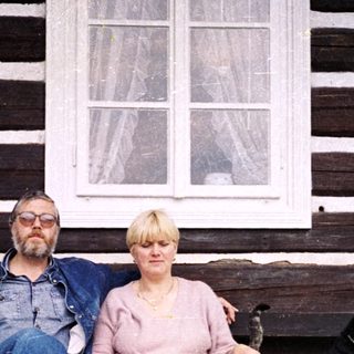 Two people sitting on bench outside rustic wooden house with pets.