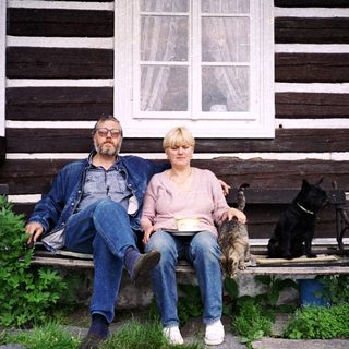 Two people sitting on bench outside rustic wooden house with pets.