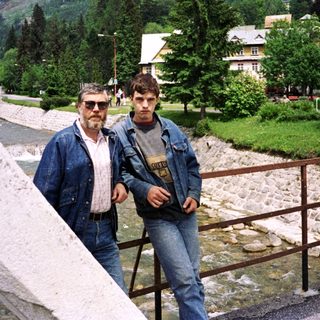 Two men posing by a scenic riverside in a mountainous area.