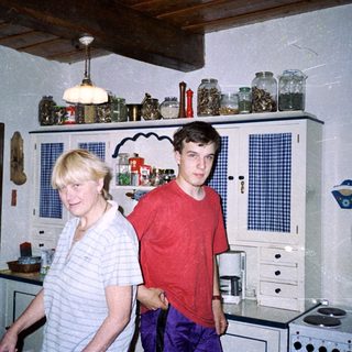 Two people standing in a vintage kitchen with wooden cabinets.