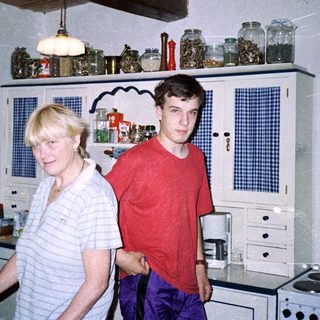 Two people standing in a vintage kitchen with wooden cabinets.