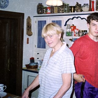 Two people standing in a vintage kitchen with wooden cabinets.