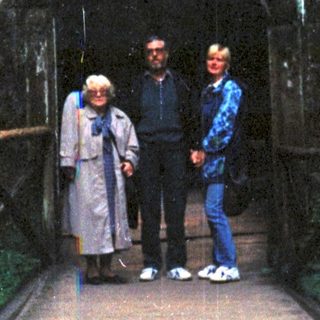 Three people standing on a wooden bridge leading into a dark cave entrance.