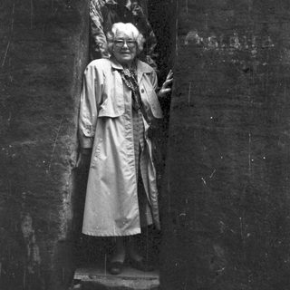 Statue of elderly woman in coat, posing outdoors.