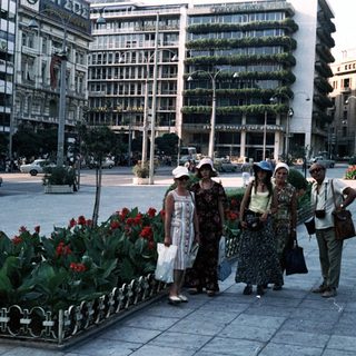 People walking in a city square with vintage cars and floral planters.